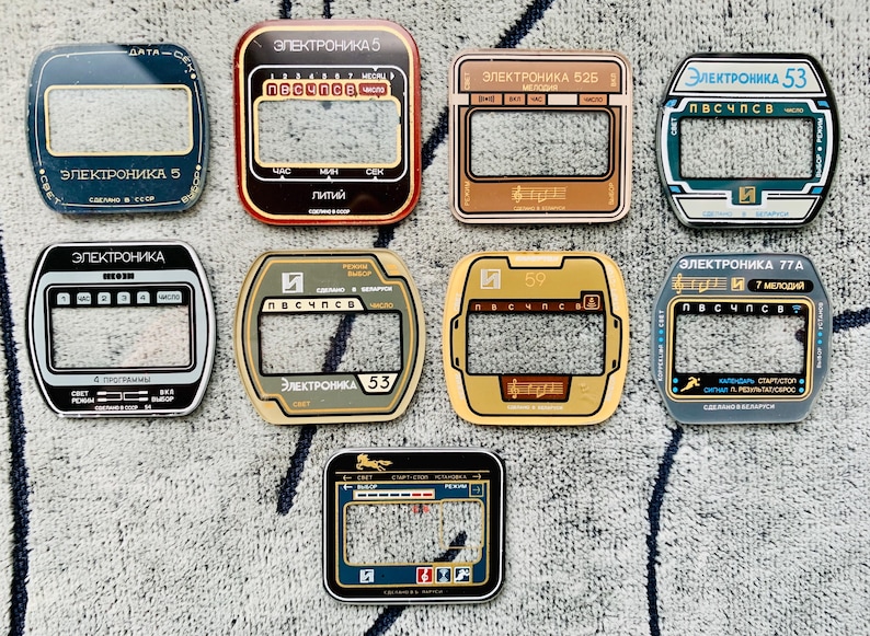May include: A collection of vintage electronic watch faceplates in various colors, including black, gold, and blue. Each plate features a rectangular display window and Cyrillic text, with designs that include musical notes and numbers.