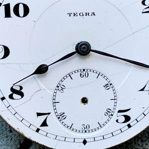 Puede incluir: Primer plano de una esfera de reloj vintage con la marca "TEGRA" en la parte superior. La esfera blanca presenta n&uacute;meros negros, un dial de segundos m&aacute;s peque&ntilde;o y manecillas negras. La esfera tiene grietas visibles, lo que aumenta su encanto antiguo.