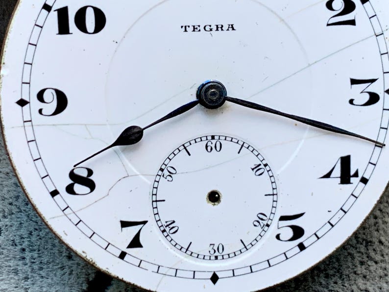 Puede incluir: Primer plano de una esfera de reloj vintage con la marca "TEGRA" en la parte superior. La esfera blanca presenta n&uacute;meros negros, un dial de segundos m&aacute;s peque&ntilde;o y manecillas negras. La esfera tiene grietas visibles, lo que aumenta su encanto antiguo.