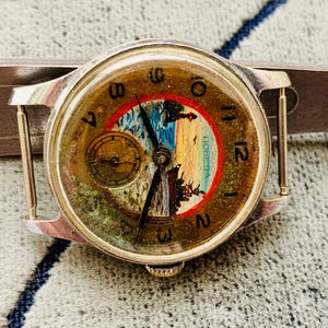 Puede incluir: Reloj de pulsera vintage con caja plateada y esfera redonda. La esfera del reloj presenta una pintura de paisaje con un borde rojo y azul, n&uacute;meros dorados y una peque&ntilde;a subesfera. El reloj est&aacute; sujeto a una correa de metal plateado.