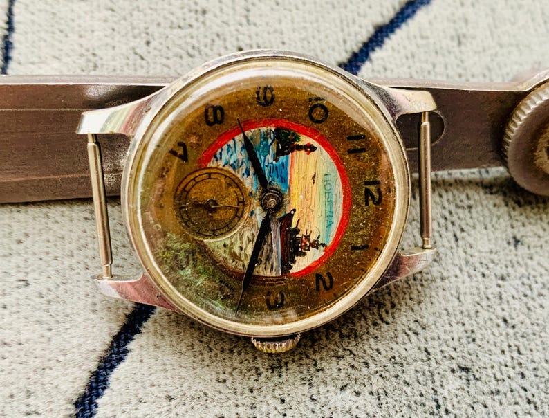 Puede incluir: Reloj de pulsera vintage con caja plateada y esfera redonda. La esfera del reloj presenta una pintura de paisaje con un borde rojo y azul, n&uacute;meros dorados y una peque&ntilde;a subesfera. El reloj est&aacute; sujeto a una correa de metal plateado.