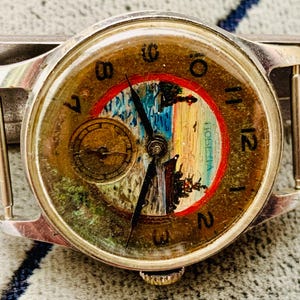 Puede incluir: Reloj de pulsera vintage con caja plateada y esfera redonda. La esfera del reloj presenta una pintura de paisaje con un borde rojo y azul, manecillas doradas y una peque&ntilde;a subesfera. El reloj tiene un aspecto texturizado y envejecido.