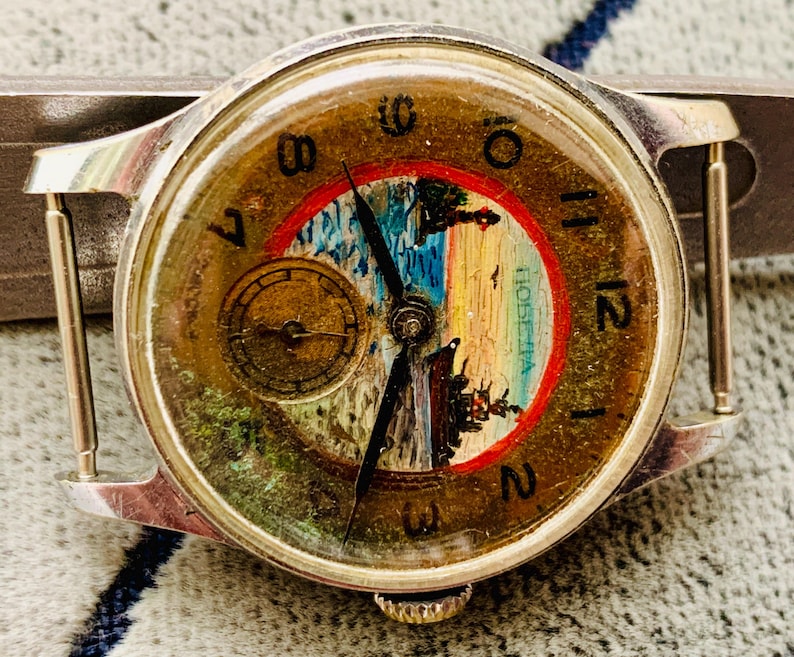Puede incluir: Reloj de pulsera vintage con caja plateada y esfera redonda. La esfera del reloj presenta una pintura de paisaje con un borde rojo y azul, manecillas doradas y una peque&ntilde;a subesfera. El reloj tiene un aspecto texturizado y envejecido.