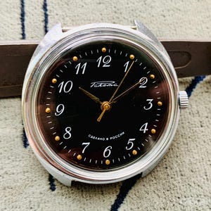 Puede incluir: Un reloj de pulsera plateado con una esfera negra y marcadores de hora de color dorado. La esfera del reloj tiene el texto "Takema" y "Сделано в России" impreso en él.