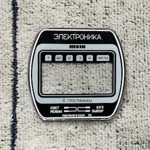 May include: A black and silver electronic device faceplate with Cyrillic text. The faceplate has a rectangular shape with rounded corners and features several buttons and a digital display area. The text includes "ЭЛЕКТРОНИКА" and "СДЕЛАНО В СССР 54".