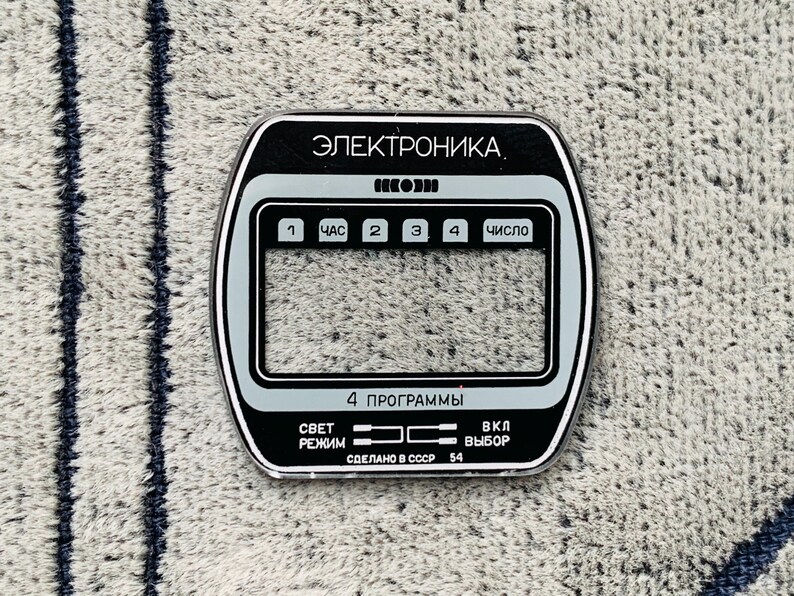 May include: A black and silver electronic device faceplate with Cyrillic text. The faceplate has a rectangular shape with rounded corners and features several buttons and a digital display area. The text includes "ЭЛЕКТРОНИКА" and "СДЕЛАНО В СССР 54".
