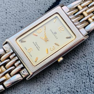 May include: A rectangular Perucci watch with a silver and gold band. The watch face is cream-colored with gold numerals and hands. The watch is marked "Expression Water Proof" and "Japan".