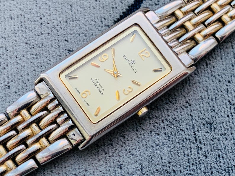 May include: A rectangular Perucci watch with a silver and gold band. The watch face is cream-colored with gold numerals and hands. The watch is marked "Expression Water Proof" and "Japan".
