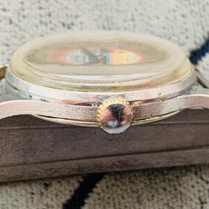 Puede incluir: Primer plano de un reloj de pulsera vintage con caja plateada y corona dorada. La esfera del reloj es visible a trav&eacute;s de un cristal transparente, ligeramente turbio. El reloj est&aacute; sujeto a una correa de cuero marr&oacute;n desgastada.