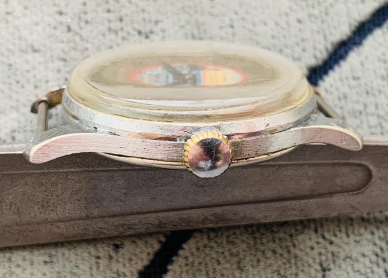 Puede incluir: Primer plano de un reloj de pulsera vintage con caja plateada y corona dorada. La esfera del reloj es visible a trav&eacute;s de un cristal transparente, ligeramente turbio. El reloj est&aacute; sujeto a una correa de cuero marr&oacute;n desgastada.