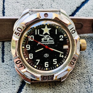 May include: A silver-toned wristwatch with a black face featuring white numerals, a star, and a tank graphic. The watch has a gold-colored crown and a red second hand. The bezel displays minute markers.
