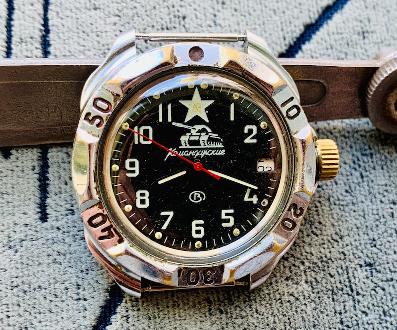 May include: A silver-toned wristwatch with a black face featuring white numerals, a star, and a tank graphic. The watch has a gold-colored crown and a red second hand. The bezel displays minute markers.