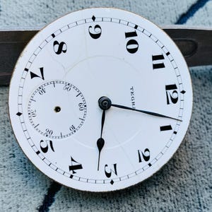 Puede incluir: Una esfera de reloj blanca con n&uacute;meros y manecillas negras. Una esfera secundaria m&aacute;s peque&ntilde;a est&aacute; en la parte inferior izquierda. La esfera del reloj tiene la palabra "TEGR" impresa. La esfera del reloj est&aacute; sobre un fondo texturizado.