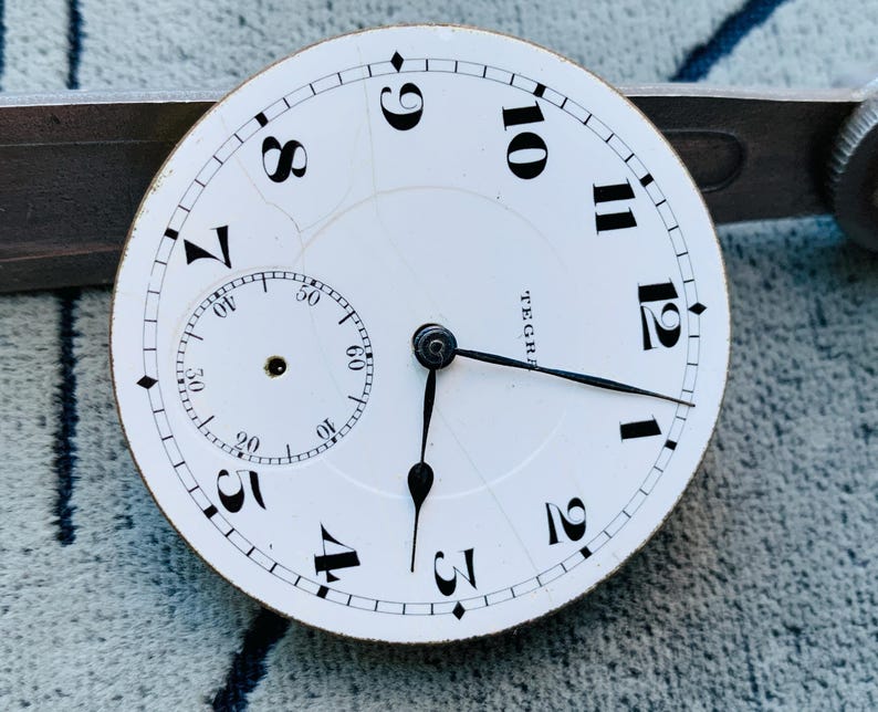 Puede incluir: Una esfera de reloj blanca con n&uacute;meros y manecillas negras. Una esfera secundaria m&aacute;s peque&ntilde;a est&aacute; en la parte inferior izquierda. La esfera del reloj tiene la palabra "TEGR" impresa. La esfera del reloj est&aacute; sobre un fondo texturizado.