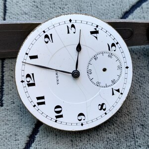Puede incluir: Una esfera de reloj de bolsillo vintage con fondo blanco y n&uacute;meros negros. La esfera del reloj tiene un peque&ntilde;o dial de segundos y agujas negras. La palabra "TEGRA" est&aacute; impresa en la esfera. La esfera del reloj est&aacute; colocada contra una herramienta de metal.