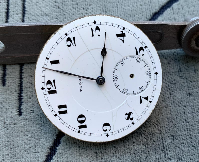 Puede incluir: Una esfera de reloj de bolsillo vintage con fondo blanco y n&uacute;meros negros. La esfera del reloj tiene un peque&ntilde;o dial de segundos y agujas negras. La palabra "TEGRA" est&aacute; impresa en la esfera. La esfera del reloj est&aacute; colocada contra una herramienta de metal.
