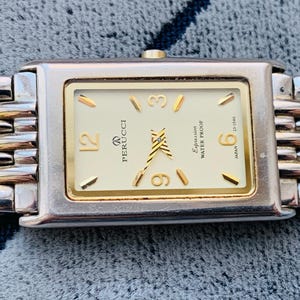 May include: A rectangular Perucci watch with a gold-tone face and silver and gold-tone band. The watch face has gold-tone hour markers and hands. The watch band is made of interlocking links. The watch face reads "Expression Water Proof Japan".