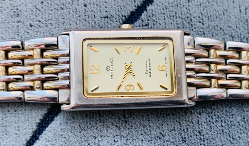 May include: A rectangular Perucci watch with a gold-tone face and silver and gold-tone band. The watch face has gold-tone hour markers and hands. The watch band is made of interlocking links. The watch face reads "Expression Water Proof Japan".