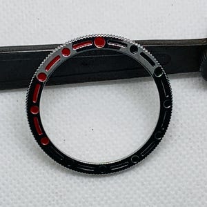 Op de afbeelding: Een zwarte en zilveren horloge bezel met rode markeringen. De bezel is een ronde ring met een glad, gepolijst oppervlak.