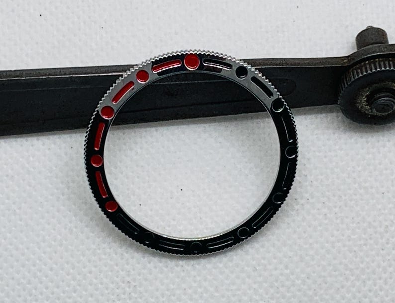 Op de afbeelding: Een zwarte en zilveren horloge bezel met rode markeringen. De bezel is een ronde ring met een glad, gepolijst oppervlak.