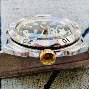May include: Close-up of a vintage wristwatch with a black face, white numerals, and a red second hand. The watch has a silver bezel with the numbers 01 and 02. The crown is gold-colored.