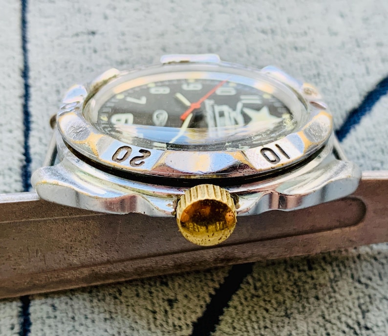 May include: Close-up of a vintage wristwatch with a black face, white numerals, and a red second hand. The watch has a silver bezel with the numbers 01 and 02. The crown is gold-colored.