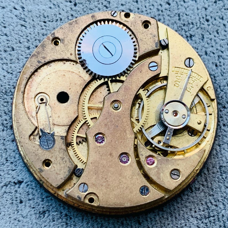 Puede incluir: Primer plano de un movimiento de reloj antiguo, que muestra intrincados engranajes, resortes y un volante. Los componentes de color lat&oacute;n est&aacute;n dispuestos en un patr&oacute;n circular, con un engranaje azul claro en la parte superior. Peque&ntilde;as joyas a&ntilde;aden un toque de color.