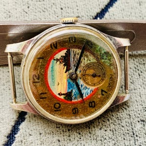 Puede incluir: Reloj de pulsera plateado vintage con esfera redonda que muestra una escena de paisaje dentro de un c&iacute;rculo rojo. El reloj tiene agujas negras, n&uacute;meros dorados y una peque&ntilde;a subesfera. El reloj est&aacute; siendo medido por una herramienta plateada.