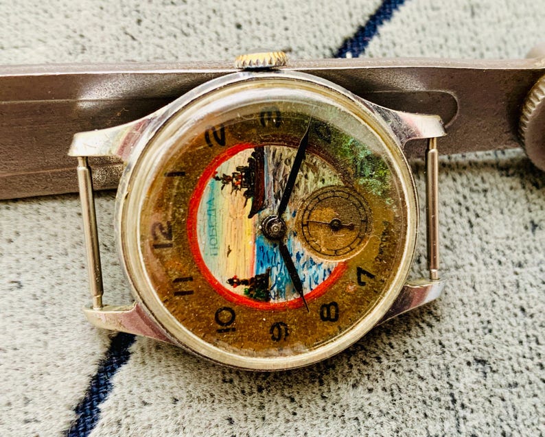 Puede incluir: Reloj de pulsera plateado vintage con esfera redonda que muestra una escena de paisaje dentro de un c&iacute;rculo rojo. El reloj tiene agujas negras, n&uacute;meros dorados y una peque&ntilde;a subesfera. El reloj est&aacute; siendo medido por una herramienta plateada.
