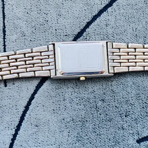 May include: A silver-toned rectangular wristwatch with a brick-patterned band. The watch face is a simple rectangle. The band is made of small, rectangular links. The watch is displayed on a textured, blue-gray surface.