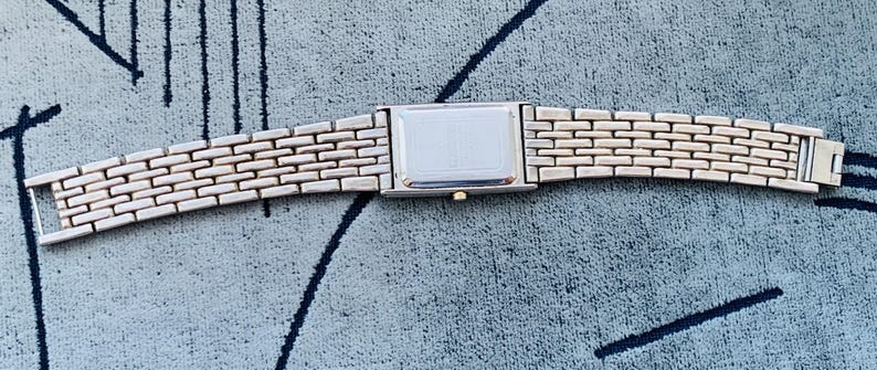 May include: A silver-toned rectangular wristwatch with a brick-patterned band. The watch face is a simple rectangle. The band is made of small, rectangular links. The watch is displayed on a textured, blue-gray surface.