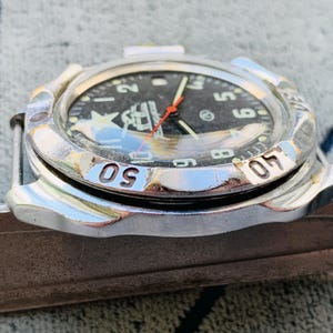 May include: A silver-toned wristwatch with a black face, featuring white numerals and a red second hand. The watch has a rotating bezel with numbers and is set on a brown surface. The watch face displays a logo.