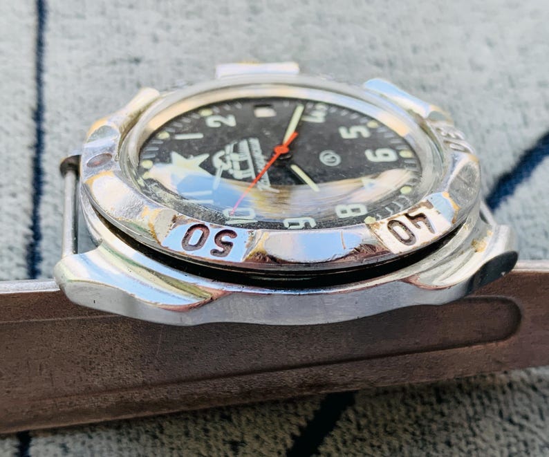 May include: A silver-toned wristwatch with a black face, featuring white numerals and a red second hand. The watch has a rotating bezel with numbers and is set on a brown surface. The watch face displays a logo.