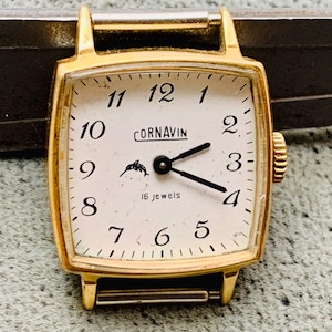 May include: A gold-tone wristwatch with a white face and black hands. The watch face has black Roman numeral hour markers and a black dolphin logo. The watch face also says "Cornavin" and "16 Jewels".