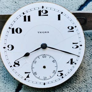 Puede incluir: Una esfera de reloj blanca con n&uacute;meros y manecillas negras. La esfera del reloj tiene una subesfera para los segundos y la marca "TEGRA". La esfera del reloj est&aacute; agrietada y muestra signos de edad.