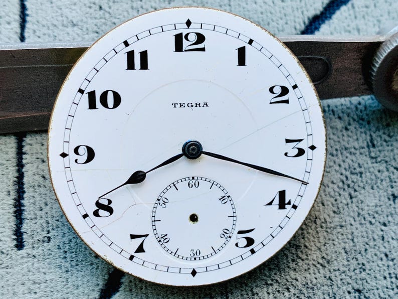 Puede incluir: Una esfera de reloj blanca con n&uacute;meros y manecillas negras. La esfera del reloj tiene una subesfera para los segundos y la marca "TEGRA". La esfera del reloj est&aacute; agrietada y muestra signos de edad.