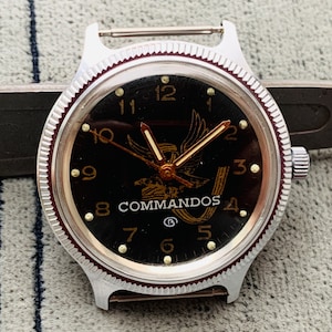 May include: A black watch face with gold accents and a gold eagle design. The watch face reads "COMMANDOS" and has numbers 1 through 12 around the edge. The watch has a silver band.