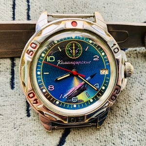 Op de afbeelding: Een zilverkleurig polshorloge met een blauwe wijzerplaat met een kleurrijke illustratie en het woord "Командирские". Het horloge heeft een rode secondewijzer en een datumweergave. De bezel heeft zwarte cijfers.