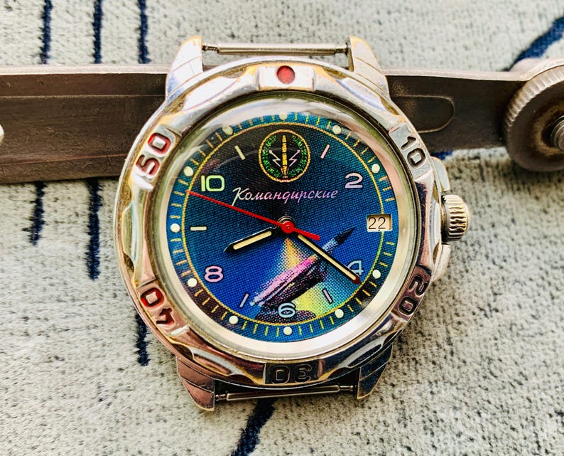 Op de afbeelding: Een zilverkleurig polshorloge met een blauwe wijzerplaat met een kleurrijke illustratie en het woord "Командирские". Het horloge heeft een rode secondewijzer en een datumweergave. De bezel heeft zwarte cijfers.