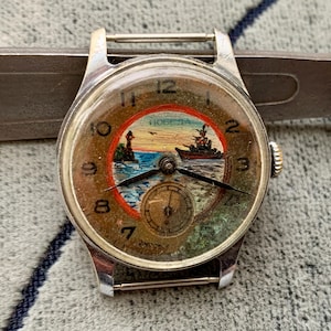Puede incluir: Reloj de pulsera plateado vintage con esfera redonda que representa una escena n&aacute;utica. La esfera muestra un barco, un faro y la palabra "Победа". El reloj tiene agujas negras y una peque&ntilde;a subesfera. Fondo gris.