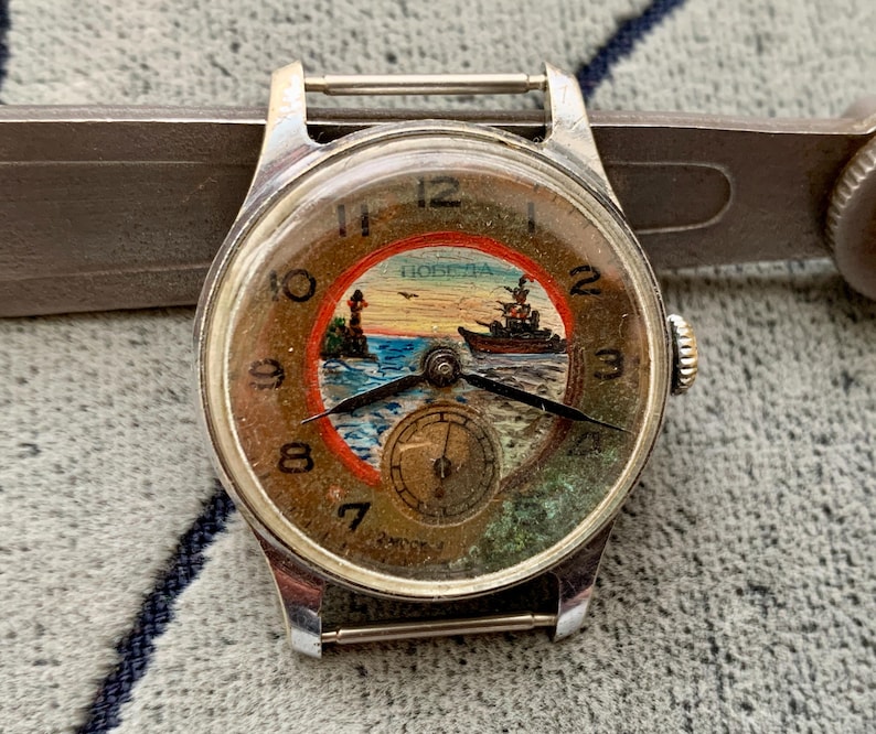 Puede incluir: Reloj de pulsera plateado vintage con esfera redonda que representa una escena n&aacute;utica. La esfera muestra un barco, un faro y la palabra "Победа". El reloj tiene agujas negras y una peque&ntilde;a subesfera. Fondo gris.
