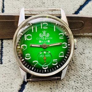 May include: A vintage CORNAVIN de luxe wristwatch with a vibrant green face. The watch features white numerals, silver hands, and the text "23 jewels." The watch is set against a silver metal band.