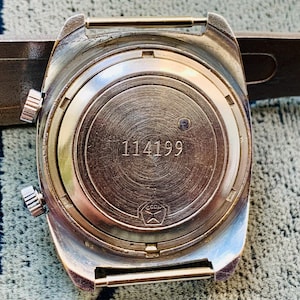 Puede incluir: Primer plano de la parte trasera de un reloj de pulsera vintage. El reloj tiene una caja de color plateado con dos botones laterales y una corona. La esfera del reloj es de metal cepillado con el n&uacute;mero "114199" grabado. Un s&iacute;mbolo es visible en la parte inferior.
