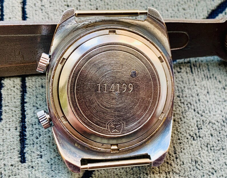 Puede incluir: Primer plano de la parte trasera de un reloj de pulsera vintage. El reloj tiene una caja de color plateado con dos botones laterales y una corona. La esfera del reloj es de metal cepillado con el n&uacute;mero "114199" grabado. Un s&iacute;mbolo es visible en la parte inferior.