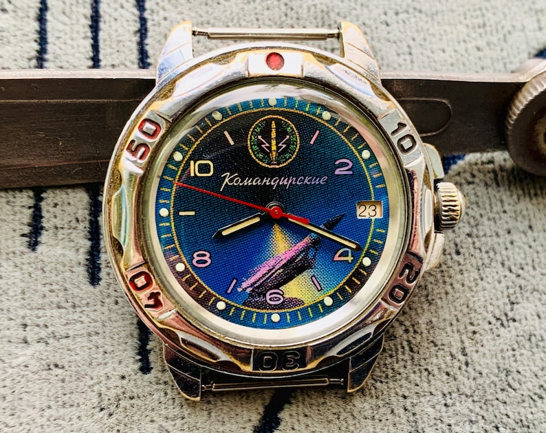 Op de afbeelding: Een zilverkleurig polshorloge met een blauwe en gele wijzerplaat. De wijzerplaat heeft de tekst "Командирские" en een afbeelding van een vliegtuig. Het horloge heeft een rode secondewijzer en een datumweergave op 3 uur.