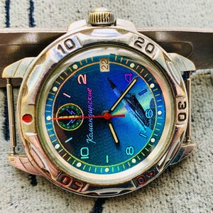Op de afbeelding: Een zilverkleurig polshorloge met een blauw en groen iriserende wijzerplaat. Het horloge heeft zwarte wijzers, een rode secondewijzer en witte cijfers. De wijzerplaat heeft de tekst "Командирские". Het horloge ligt op een grijze achtergrond.