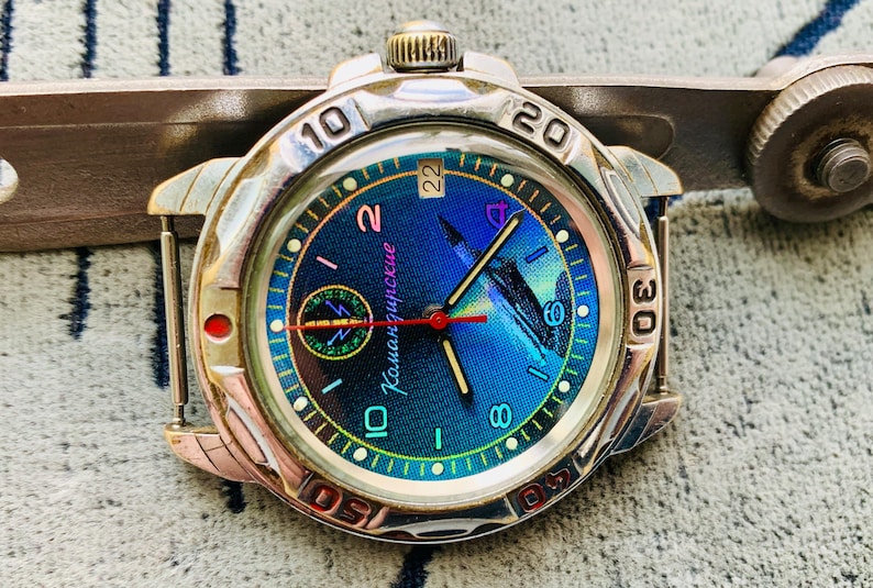 Op de afbeelding: Een zilverkleurig polshorloge met een blauw en groen iriserende wijzerplaat. Het horloge heeft zwarte wijzers, een rode secondewijzer en witte cijfers. De wijzerplaat heeft de tekst "Командирские". Het horloge ligt op een grijze achtergrond.