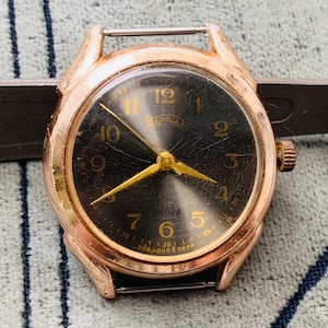 May include: A gold-toned wristwatch with a black face and gold-coloured hour and minute hands. The watch has a black leather strap and a white dial with black numbers.