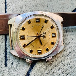 Puede incluir: Un reloj de pulsera vintage Poljot 18 jewels con esfera dorada, manecillas negras y caja plateada. La esfera del reloj muestra marcadores de hora rectangulares negros y los n&uacute;meros 3, 6, 9 y 12. El reloj est&aacute; sobre un fondo texturizado.
