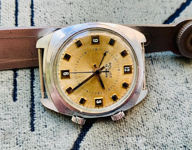 Puede incluir: Un reloj de pulsera vintage Poljot 18 jewels con esfera dorada, manecillas negras y caja plateada. La esfera del reloj muestra marcadores de hora rectangulares negros y los n&uacute;meros 3, 6, 9 y 12. El reloj est&aacute; sobre un fondo texturizado.